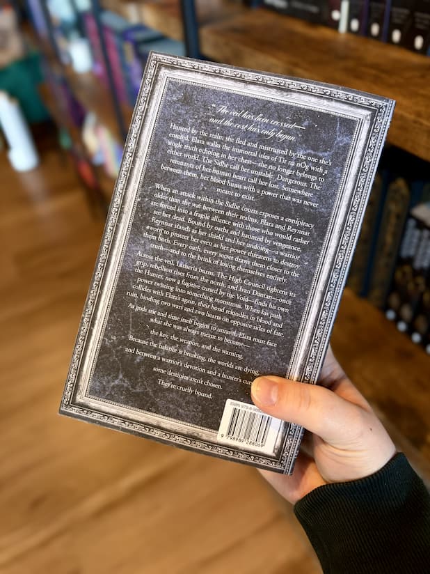 A hand holding the back of a copy of 'Fae So Cruelly Bound' by J.M. featuring a dark textured background that is framed by a silver ornate frame. 