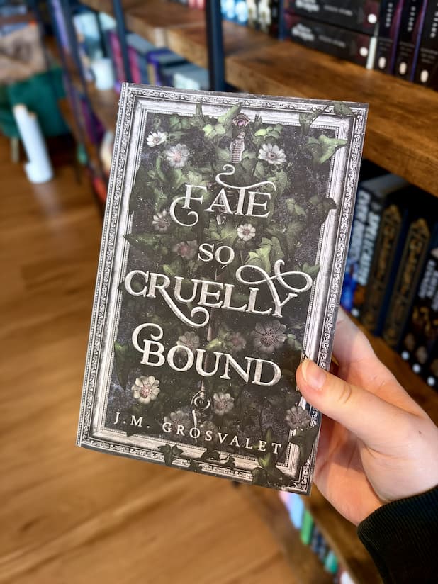 A hand holding a copy of 'Fae So Cruelly Bound' by J.M. with a silver sword laying in a bed of flowers and leaves framed by a silver ornate frame. 