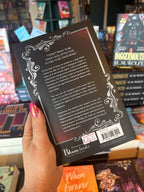 A hand holds the back cover for 'A Lady of Rooksgrave Manor' by Kathryn Moon, featuring a blurb written in white text framed by a silver ornate border, in front of a table full of blurry books.