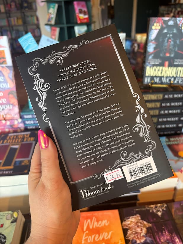A hand holds the back cover for 'A Lady of Rooksgrave Manor' by Kathryn Moon, featuring a blurb written in white text framed by a silver ornate border, in front of a table full of blurry books.