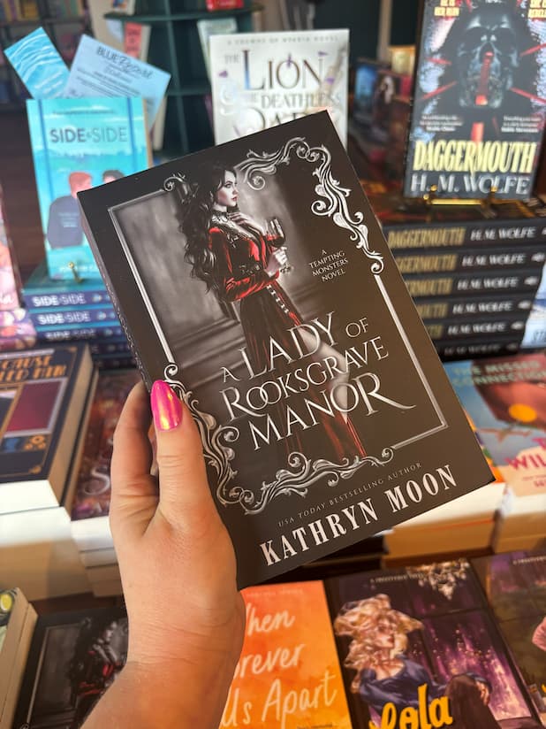 A hand holds the book cover for 'A Lady of Rooksgrave Manor' by Kathryn Moon, featuring a woman in a long red dress holding a wine glass against a dark gothic interior, framed by ornate silver scrollwork on a black background, in front of a table full of blurry books.