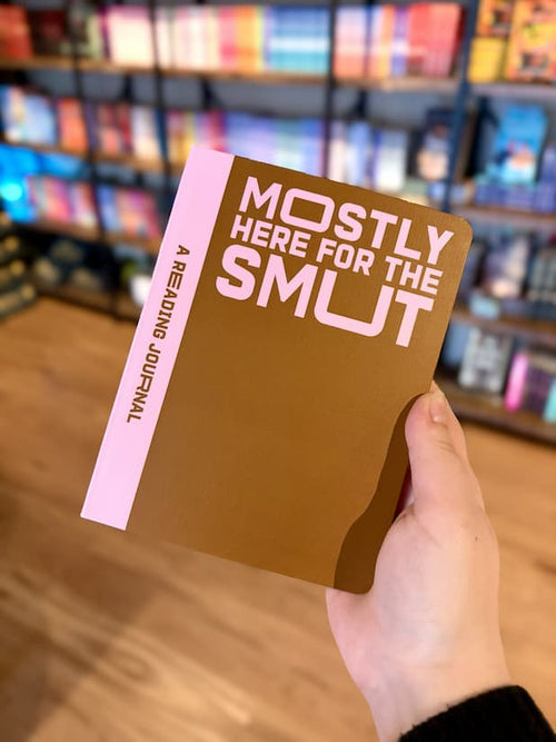A hand holding a reading journal with 'Mostly Here for the Smut' text on a pink and brown background