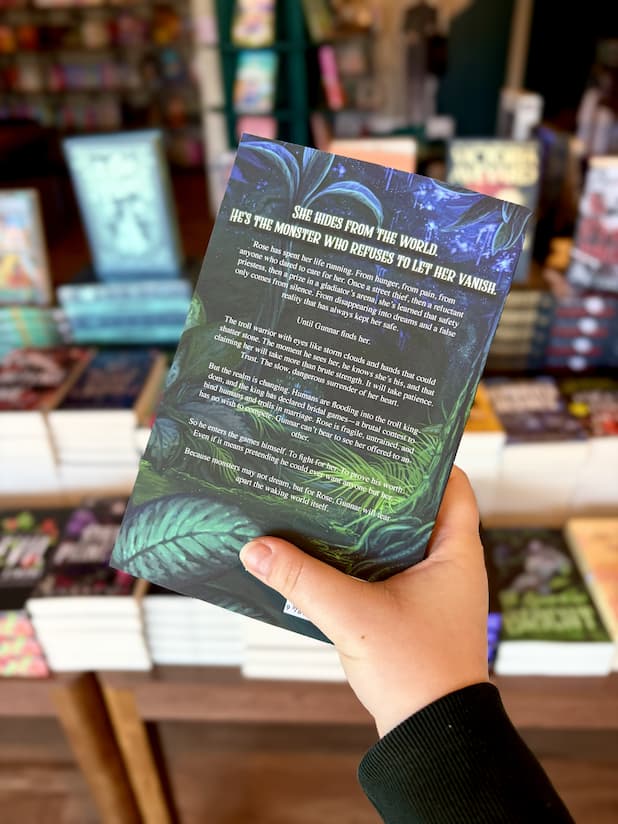 A hand holding the back of a copy of 'A Spark so Bright' featuring a blue and green fantasy forest scene with white text.
