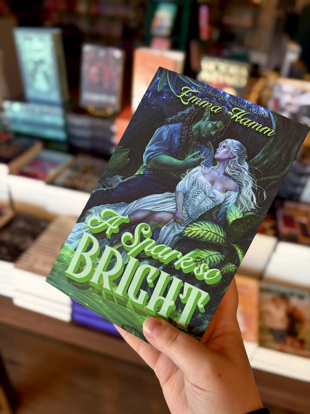 A hand holding a copy of 'A Spark so Bright' featuring a fantasy scene with a green-skinned orc caressing the blond hair of a woman in a forest.