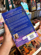 A hand holds a the back cover of 'A Spell for Midwinter's Heart' with the blurb in white font on a dark blue background, in front of a wooden table stacked with other blurry books.