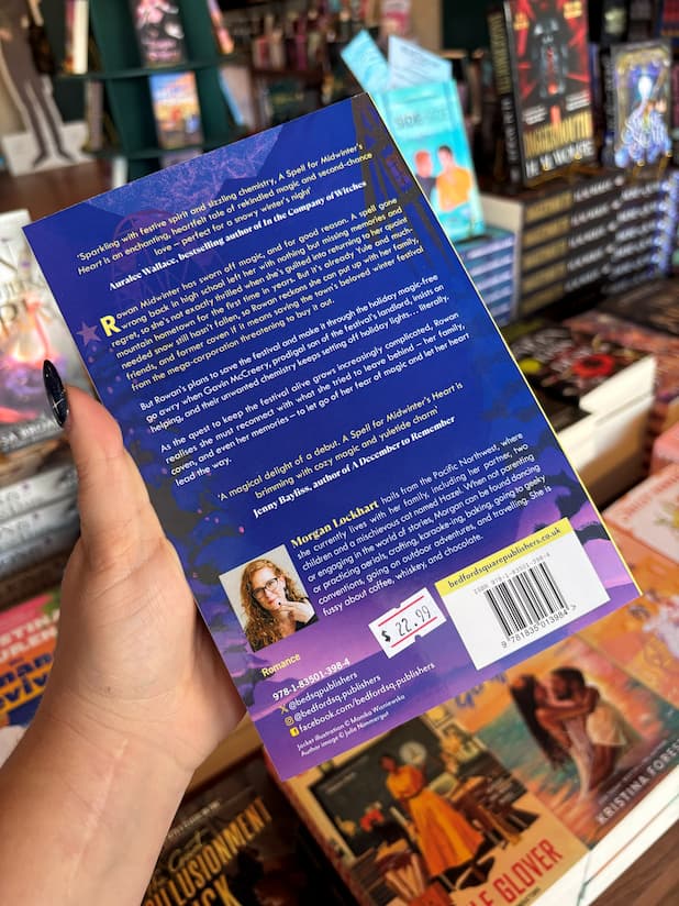 A hand holds a the back cover of 'A Spell for Midwinter's Heart' with the blurb in white font on a dark blue background, in front of a wooden table stacked with other blurry books.