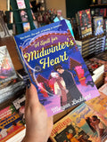 A hand holds a copy of Book cover of 'A Spell for Midwinter's Heart' with a couple in a snowy scene and a heart-shaped spell on the cover, in front of a wooden table stacked with other blurry books.
