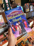 A hand holds a copy of Book cover of 'A Spell for Midwinter's Heart' with a couple in a snowy scene and a heart-shaped spell on the cover, in front of a wooden table stacked with other blurry books.