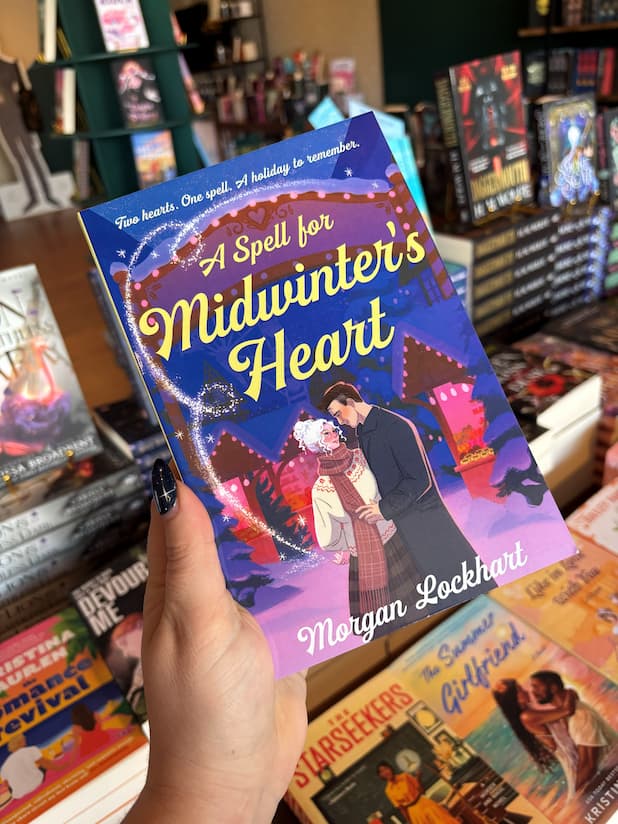 A hand holds a copy of Book cover of 'A Spell for Midwinter's Heart' with a couple in a snowy scene and a heart-shaped spell on the cover, in front of a wooden table stacked with other blurry books.