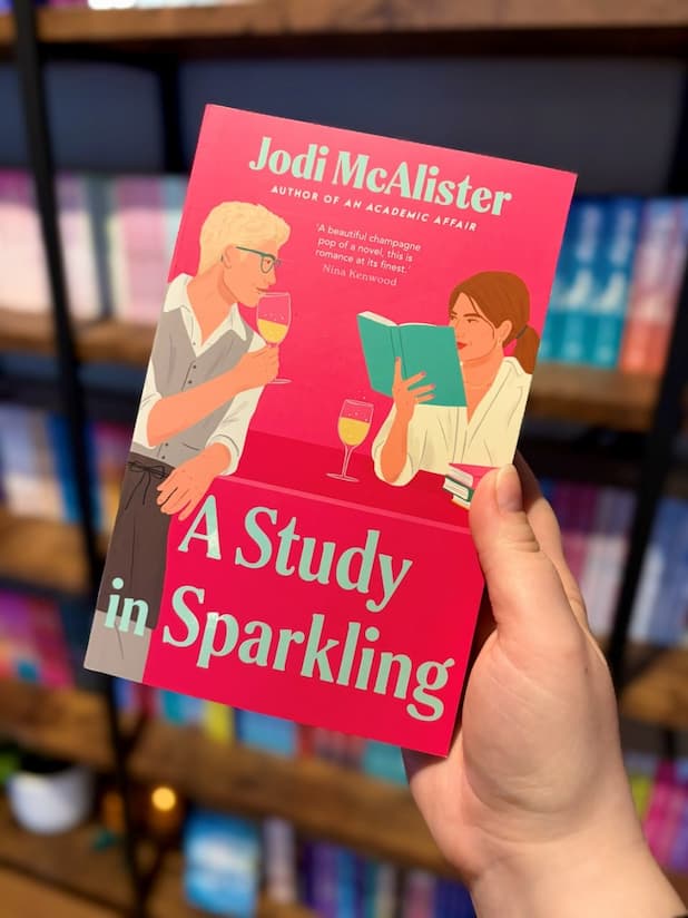 A hand holding a copy of 'A Study in Sparkling' by Jodi McAlister with illustrations of a man and a woman looking at each other across a glass of sparkling wine and a book.