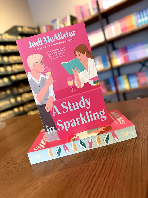 A small stack of 'A Study in Sparkling' by Jodi McAlister with illustrations of a man and a woman looking at each other across a glass of sparkling wine and a book, and sprayed edges of books.