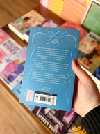 A hand holding the back cover of a blue book called A Summer of Stars by Liana Cincotti, with a blurred background of other books on a wooden surface.