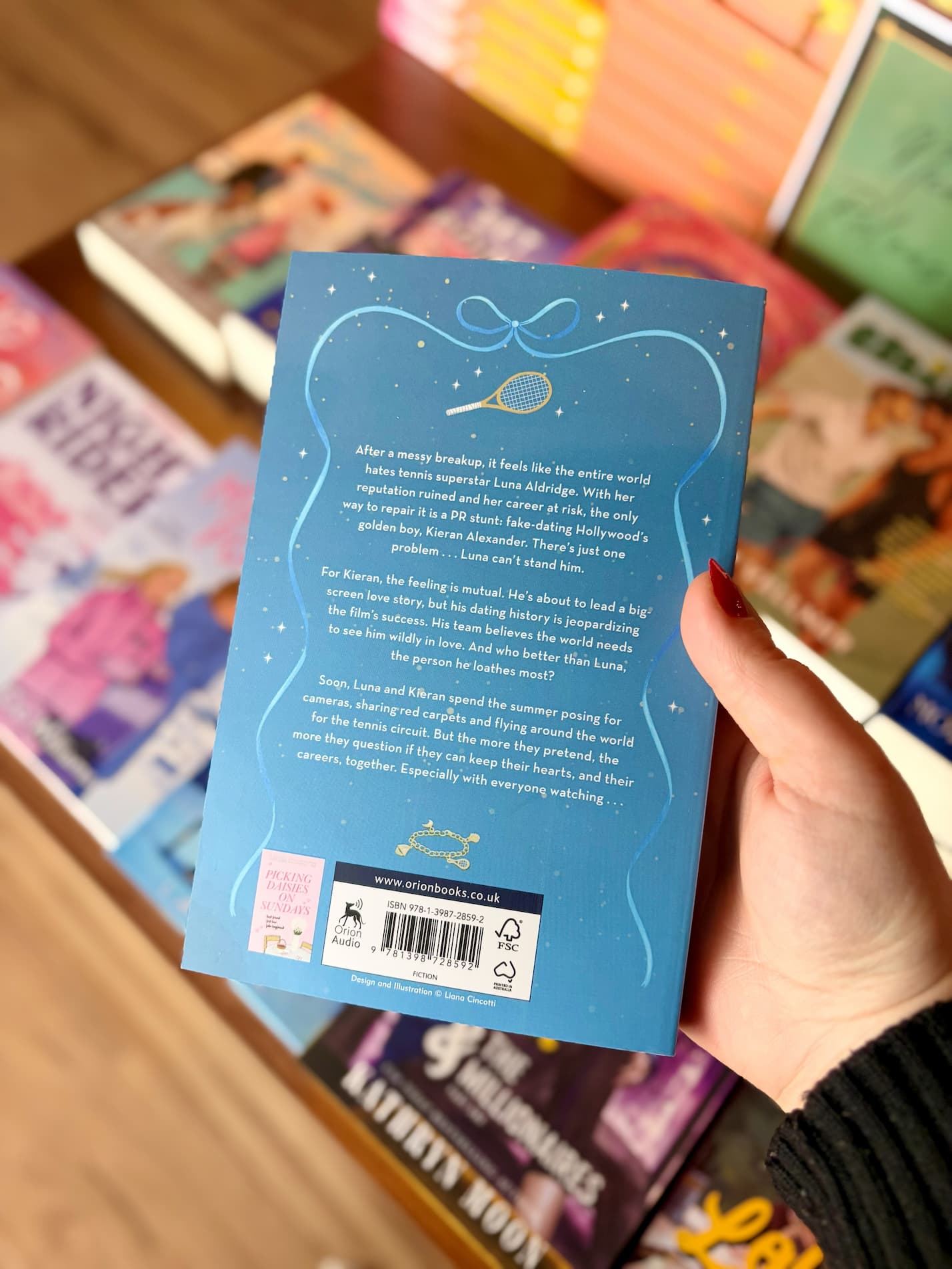 A hand holding the back cover of a blue book called A Summer of Stars by Liana Cincotti, with a blurred background of other books on a wooden surface.