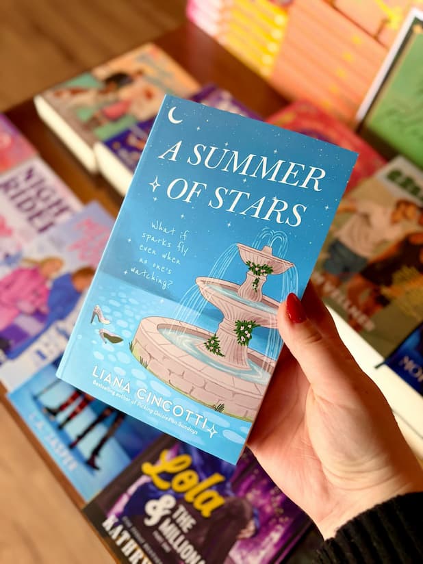 A hand with red nail polish holds a paperback book titled 'A Summer of Stars' by Liana Cincotti. The book cover features a light blue background with an illustration of a fountain and stars, and a pair of silver heels to the left of the fountain. Below the title reads a subtitle "what if sparks fly even when no one's watching"