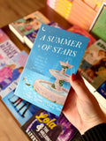 A hand with red nail polish holds a paperback book titled 'A Summer of Stars' by Liana Cincotti. The book cover features a light blue background with an illustration of a fountain and stars, and a pair of silver heels to the left of the fountain. Below the title reads a subtitle "what if sparks fly even when no one's watching"