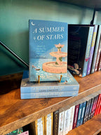 A copy of the book 'A Summer of Stars' by Liana Cincotti displayed on a wooden bookshelf, standing upright on a small gold book stand next to other copies of the book.