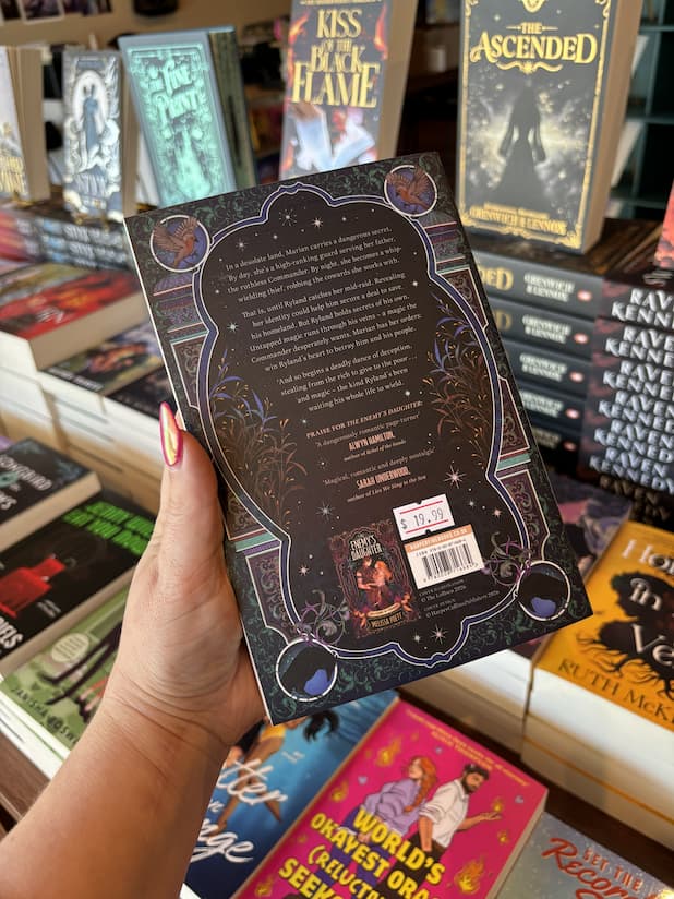 A hand with pink nail polish holding the back of a copy of 'A Thief to Die For' by Melissa Poett, featuring a black background with white text and decorative borders in blue, green and purple. 
