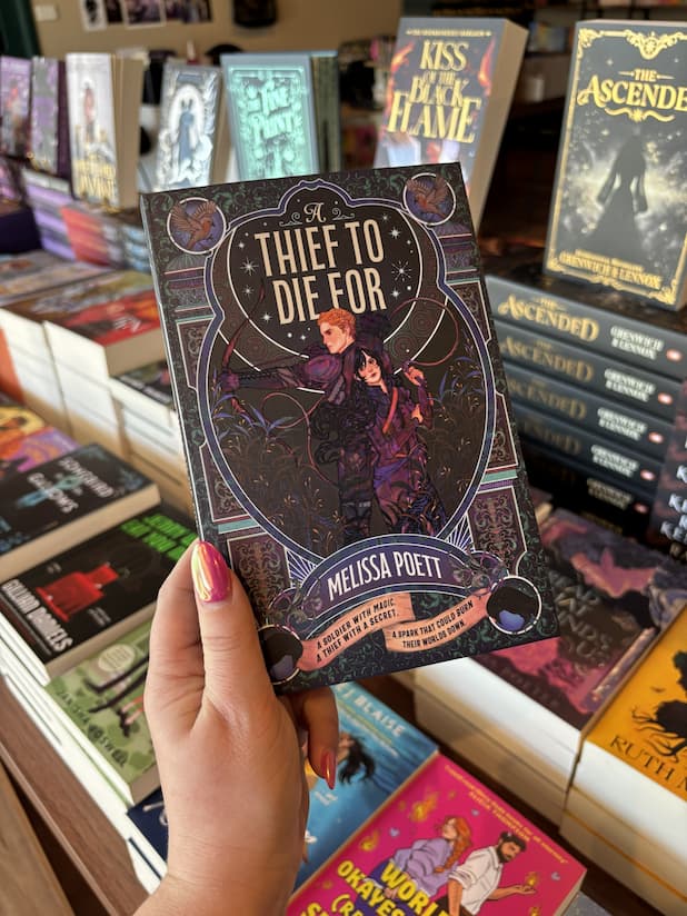 A hand with pink nail polish holding a copy of 'A Thief to Die For' by Melissa Poett, featuring a man wielding a bow and arrow standing back-to-back with a woman in a hood holding a whip. They are surrounded by decorative borders in blue, green and purple. 