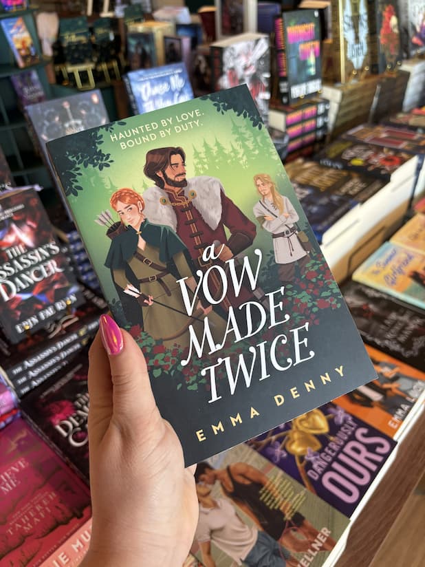 A hand with pink nail polish holding a cover of 'A Vow Made Twice' featuring three characters in a forest setting: a woman with red hair and a bow and arrow, a man in a red tunic and fur coat holding a sword at his waist, both of them are looking back at a blonde man with his arms crossed in a beige tunic and cross shoulder bag. 