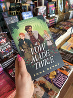 A hand with pink nail polish holding a cover of 'A Vow Made Twice' featuring three characters in a forest setting: a woman with red hair and a bow and arrow, a man in a red tunic and fur coat holding a sword at his waist, both of them are looking back at a blonde man with his arms crossed in a beige tunic and cross shoulder bag. 