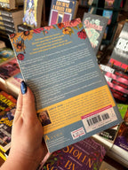 A hand holds the back cover of 'Anderson in Bloom' by Jennifer Dugan with white and yellow font for the blurb on a blue background with pink and yellow flowers framing the top of the cover, the book sits in front of a table full of blurry books in a bookstore.