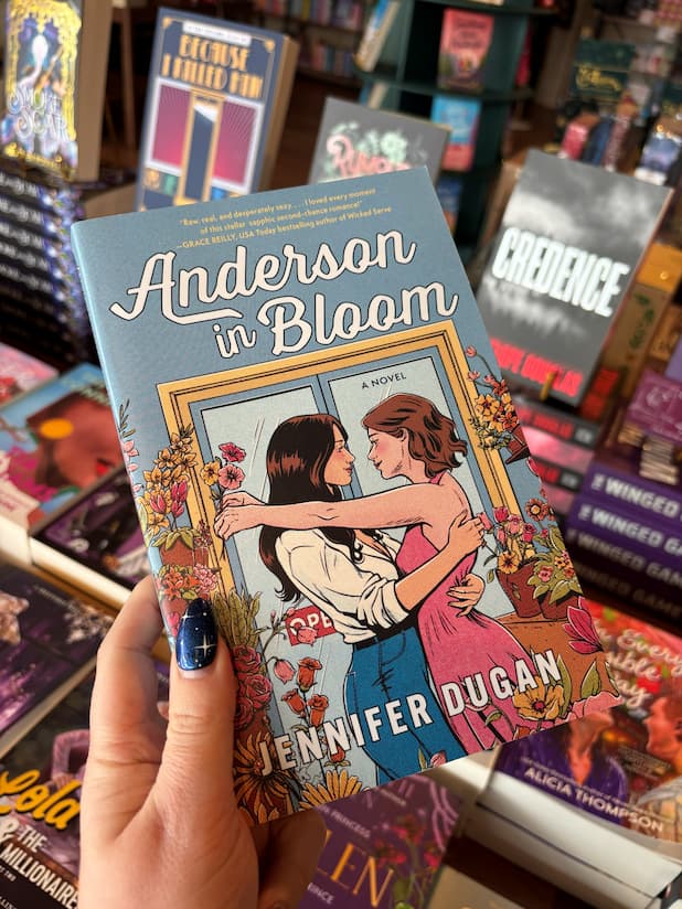 A hand holds the book cover of 'Anderson in Bloom' by Jennifer Dugan with two women embracing in a blue room filled with pink, orange and yellow flowers, the book sits in front of a table full of blurry books in a bookstore.