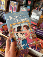 A hand holds the book cover of 'Anderson in Bloom' by Jennifer Dugan with two women embracing in a blue room filled with pink, orange and yellow flowers, the book sits in front of a table full of blurry books in a bookstore.