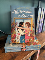 A copy of 'Anderson in Bloom' by Jennifer Dugan sits on a gold stand on top of a small stack of the same book, the cover features two women embracing in a blue room filled with pink, orange and yellow flowers, the book sits in front of a table full of blurry books in a bookstore.