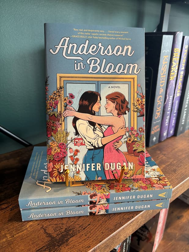 A copy of 'Anderson in Bloom' by Jennifer Dugan sits on a gold stand on top of a small stack of the same book, the cover features two women embracing in a blue room filled with pink, orange and yellow flowers, the book sits in front of a table full of blurry books in a bookstore.