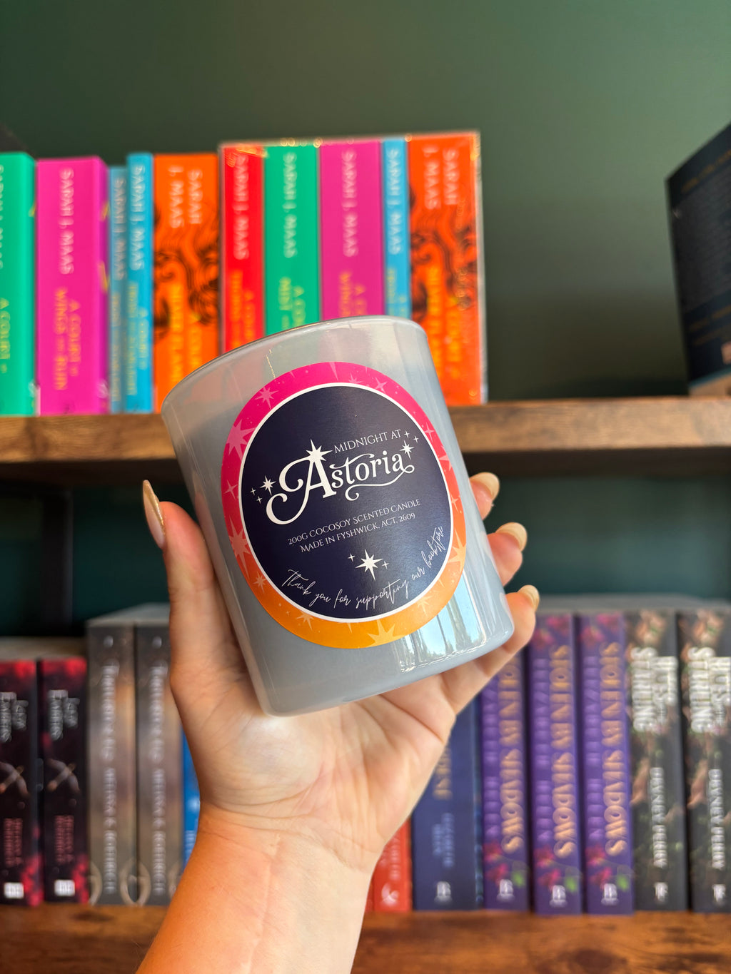 A hand holding a frosted glass candle jar with a round black, pink, and orange label that reads 'Midnight at Astoria'. The background shows a wooden bookshelf filled with colorful books against a dark green wall.