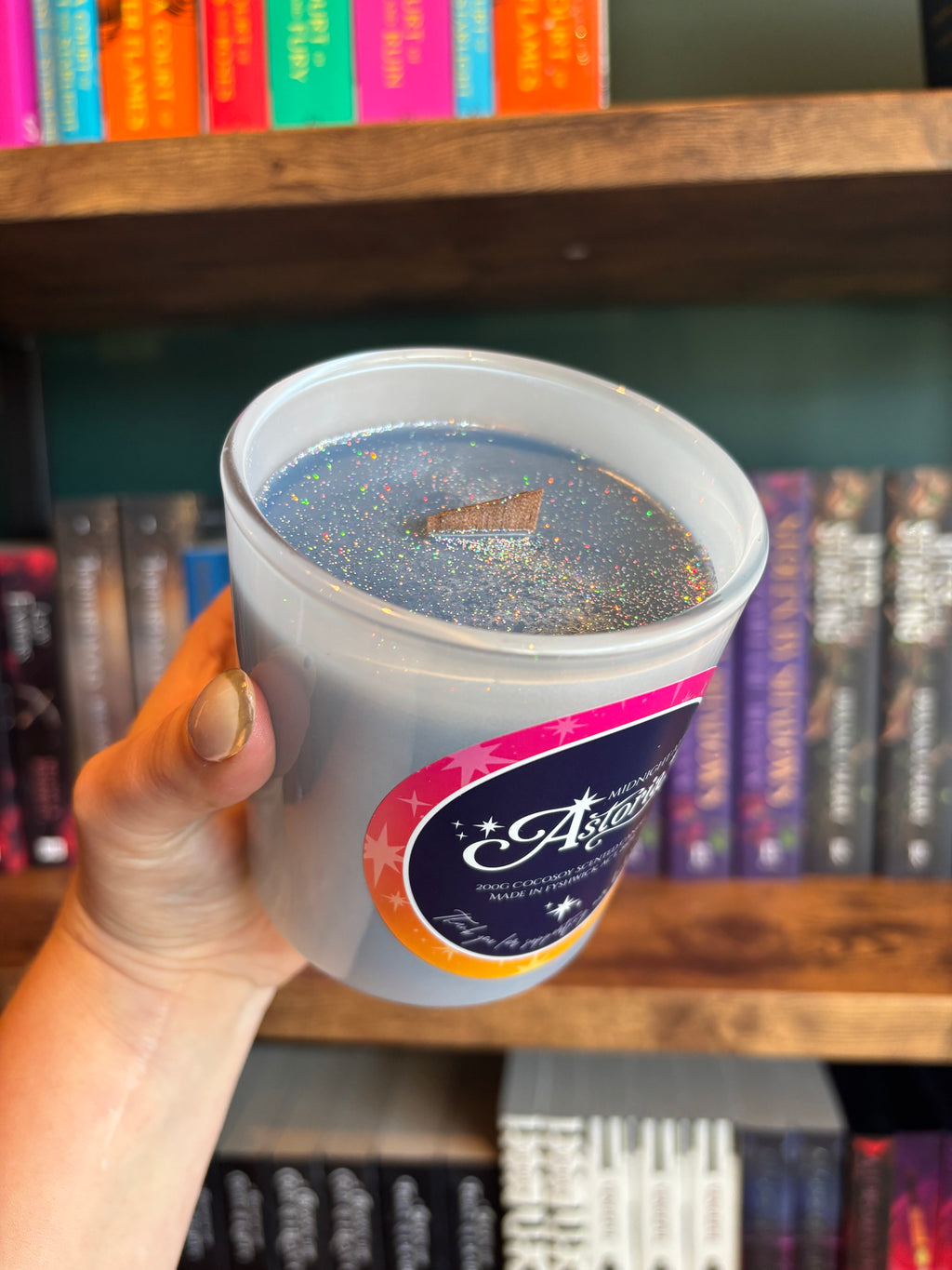 A hand holds a large white candle jar filled with blue glittery wax and a small wooden wick, with a colorful label reading 'Astoria at Midnight'. The background shows blurred wooden bookshelves filled with books.