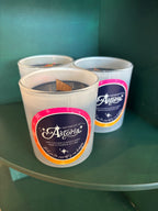 Three white soy candles in frosted glass jars with dark wax and round labels reading 'Astoria at Midnight,' displayed on a green shelf against a dark teal background.