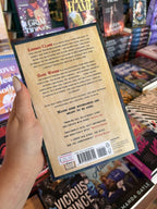A hand with nude nail polish holding the back a cover for 'Axe Marks the Spot' by Kayla Grosse, featuring a graphic wooden background and red and dark blue text.