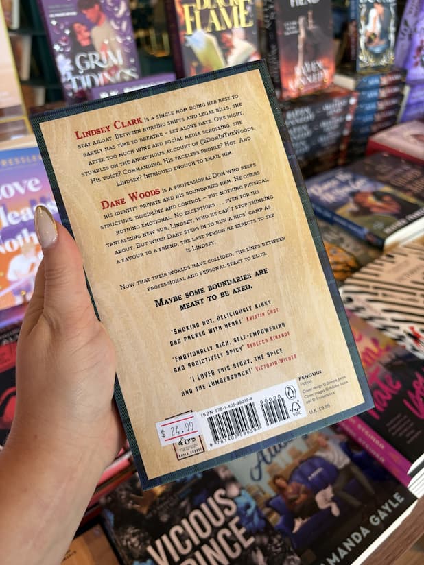 A hand with nude nail polish holding the back a cover for 'Axe Marks the Spot' by Kayla Grosse, featuring a graphic wooden background and red and dark blue text.