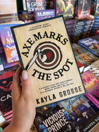 A hand with nude nail polish holding a cover for 'Axe Marks the Spot' by Kayla Grosse, featuring a graphic of a target that looks like the rings of a tree stump. The bullseye is a heart with an axe stuck through it.