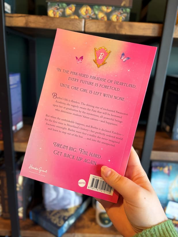 A hand holds the back cover of the book Barbie Dreamscape by Alex Aster, featuring a Barbie logo with a pink and purple butterfly on either side on top of purple text in the blurb on a pink background. A blurry bookshelf is in the background. 
