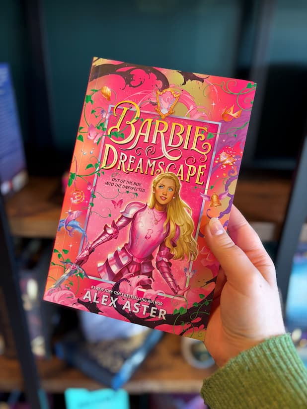 A hand holding the front cover of the book Barbie Dreamscape by Alex Aster, featuring Barbie in pink armor against a whimsical pink fantasy background with flowers, butterflies, and a sword, displayed in front of a blurry bookcase.
