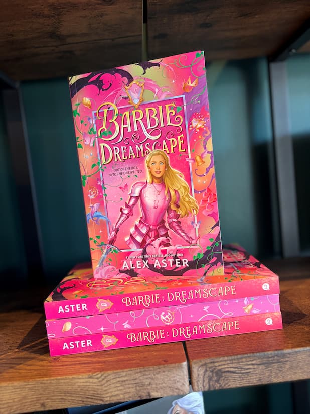 A stack of the book 'Barbie Dreamscape' by Alex Aster, alternating spine and sprayed pink edges, the cover features Barbie in pink armor against a whimsical pink fantasy background with flowers, butterflies, and a sword. The stack is displayed on a wooden bookshelf with a green background.