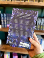 A hand holding the back of a paperback copy of 'Bedeviled' by Laura Thalassa with a mystical design showing a big tree growing up the middle with two hands reaching towards it with celestial images, snakes and birds surrounding the tree. 