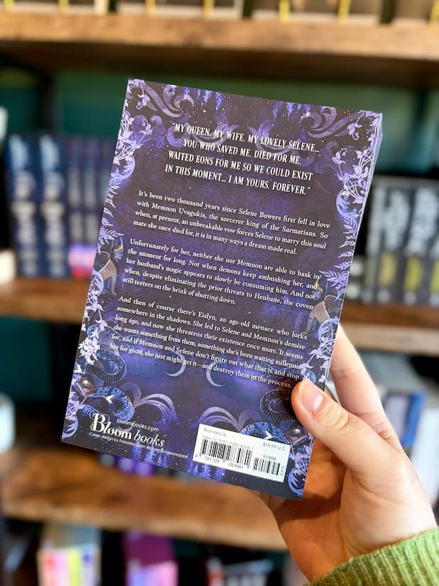 A hand holding the back of a paperback copy of 'Bedeviled' by Laura Thalassa with a mystical design showing a big tree growing up the middle with two hands reaching towards it with celestial images, snakes and birds surrounding the tree. 