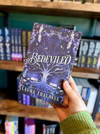 A hand holding a paperback copy of 'Bedeviled' by Laura Thalassa with a mystical design showing a big tree growing up the middle with two hands reaching towards it with celestial images, snakes and birds surrounding the tree. 