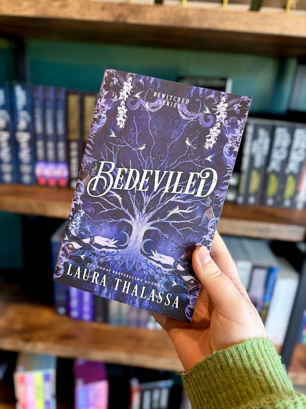 A hand holding a paperback copy of 'Bedeviled' by Laura Thalassa with a mystical design showing a big tree growing up the middle with two hands reaching towards it with celestial images, snakes and birds surrounding the tree. 