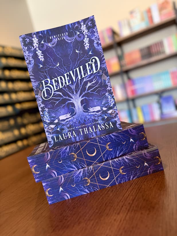 A stack of 'Bedeviled' by Laura Thalassa with a mystical design showing a big tree growing up the middle with two hands reaching towards it with celestial images, snakes and birds surrounding the tree. 