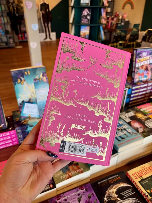 A hand holds a copy of 'Believe Me' by Tahereh Mafi, with gold foil artwork and text on the back cover in a bookstore setting, surrounded by other books on shelves.