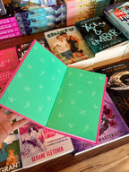 A hand holds an open copy of 'Believe Me' by Tahereh Mafi with bright green pages and pink edges, surrounded by a display of other books on a wooden shelf.