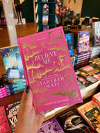 A hand holds a copy of the book 'Believe Me' by Taherah Mafi. The book has a vibrant pink cover with gold foil artwork and lettering.