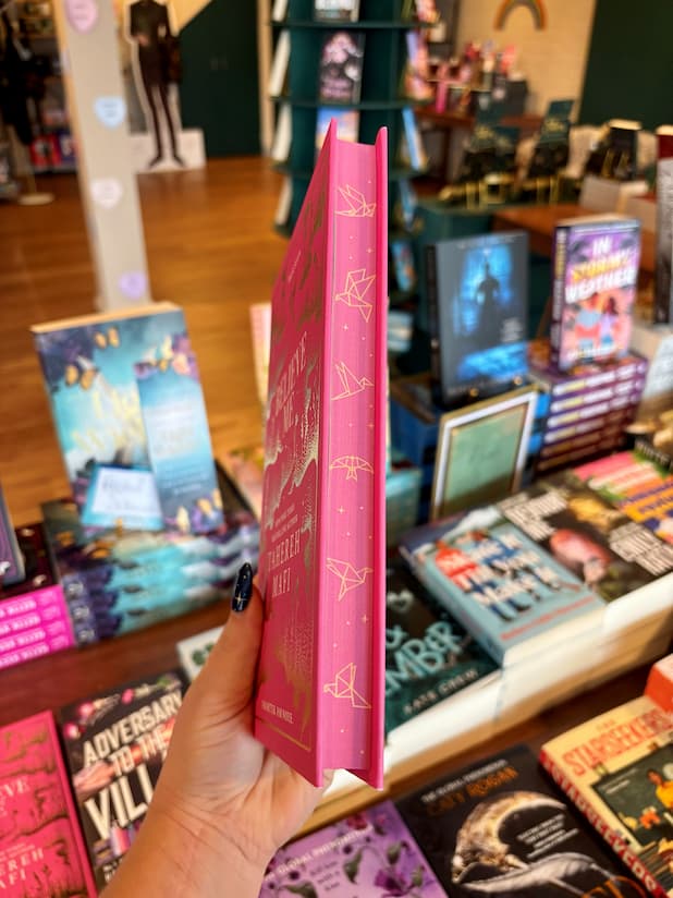 A hand holds a pink hardcover book titled 'Believe Me' by Tahereh Mafi in a bookstore, with other books displayed on shelves in the background.