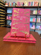 A pink hardcover book titled 'Believe Me' by Tahereh Mafi, displayed upright on a gold book stand on a wooden table, with blurred bookshelves in the background.