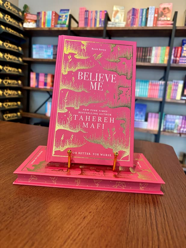 A pink hardcover book titled 'Believe Me' by Tahereh Mafi, displayed upright on a gold book stand on a wooden table, with blurred bookshelves in the background.