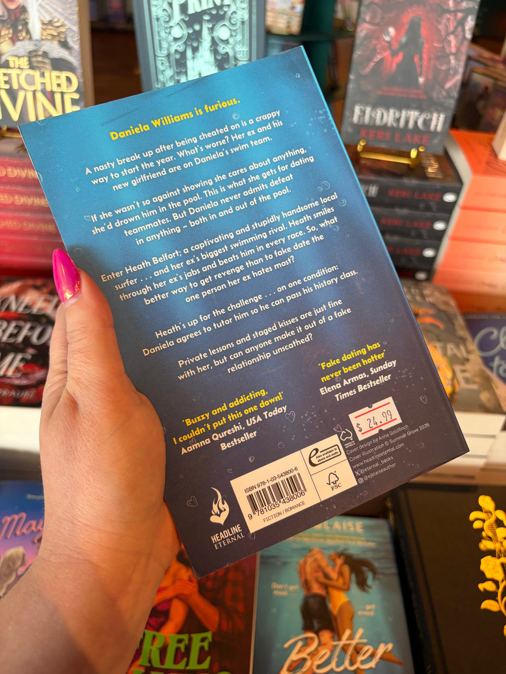 A hand with pink glitter nail polish holds the back of the book 'Better Than Revenge' by E.J. Blaise, featuring an underwater scene with yellow and white font for the blurb. The book is held up in a bookshop surrounded by other blurry books.